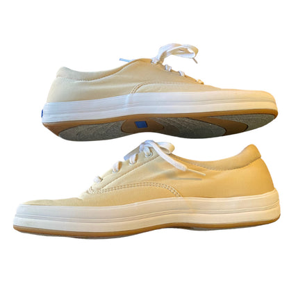 A pair of low-top canvas sneakers in a beige color, shown from the side against a plain white background. One shoe is positioned above the other, displaying the side profile, laces, and gum rubber sole.