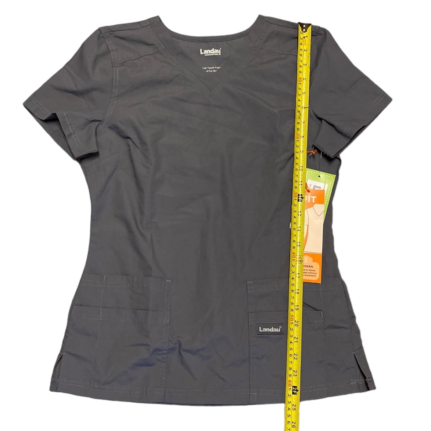 A dark gray short-sleeve scrub top , shown flat on a white background with a yellow measuring tape and retail tag standing alongside it to indicate size.