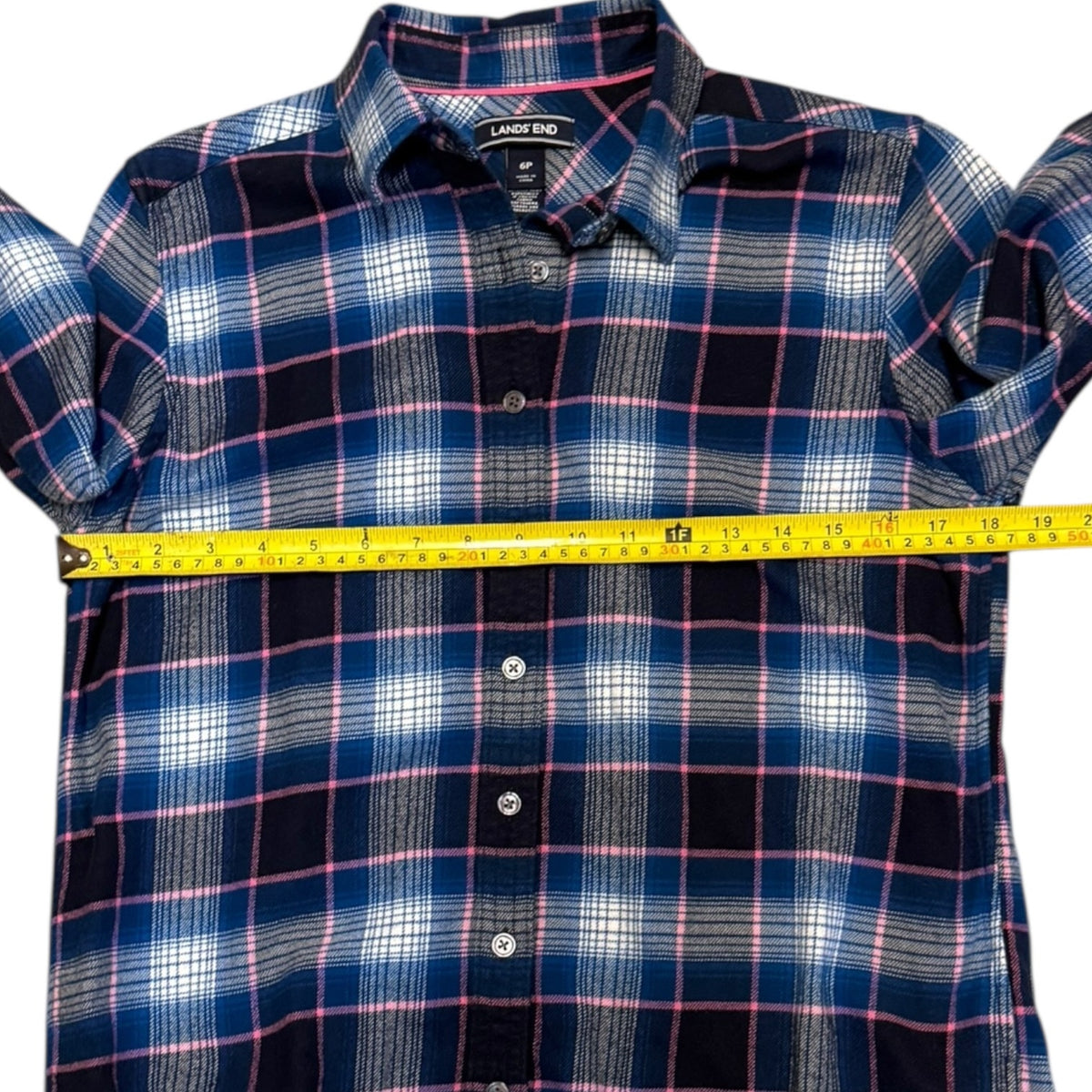 A blue plaid button-down shirt with pink and white stripes, shown with a yellow measuring tape stretched horizontally across the chest to indicate size.