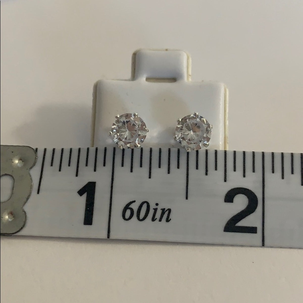 A pair of round stud earrings with clear faceted gemstones in silver-toned settings, displayed on a white card above a measuring ruler to show size.
