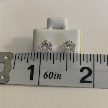 A pair of round stud earrings with clear faceted gemstones in silver-toned settings, displayed on a white card above a measuring ruler to show size.
