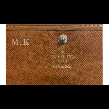 Close-up of a Louis Vuitton monogram key wallet in brown textured Saffiano canvas, featuring a silver-tone metal snap button and white printed branding text.