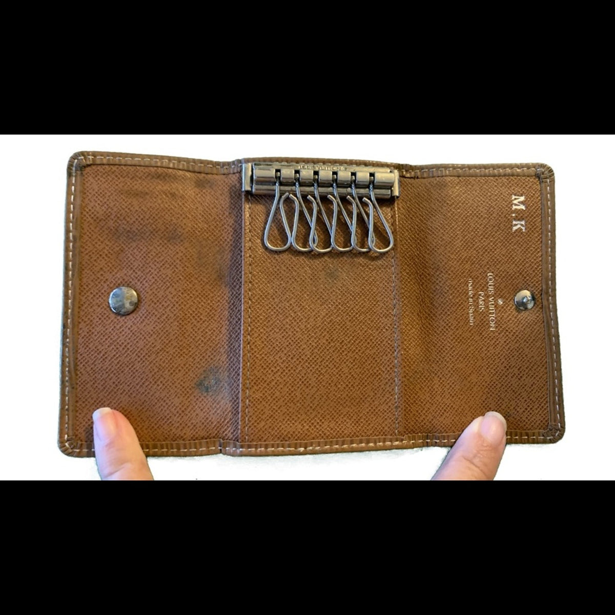 A brown textured leather keyring case held open by two fingers, showing a metal keyring with six loops, snap buttons, and small gold-toned lettering on a white background.