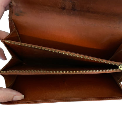 A close-up of a brown leather wallet  shown open, with a hand holding one corner to demonstrate the interior space and a zippered compartment.
