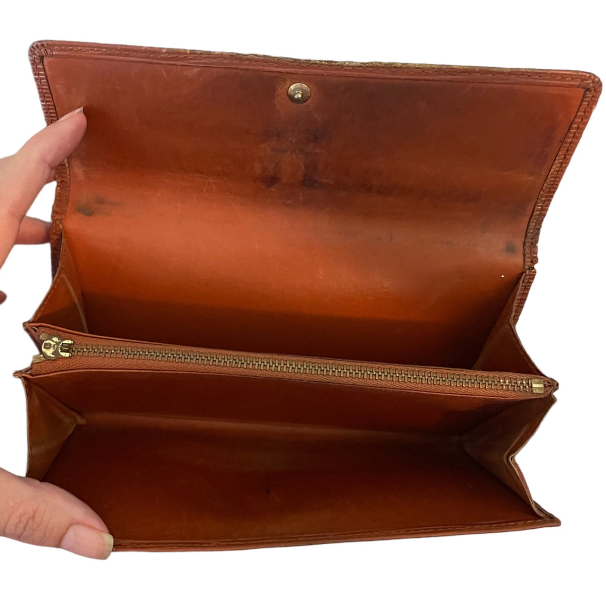 An open brown leather wallet shown against a white background, with a hand holding the left side to display the interior compartments and gold-tone zipper.