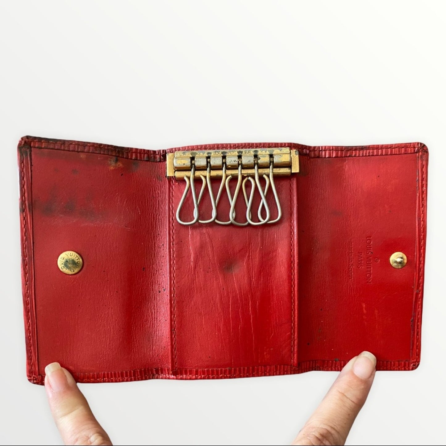 A vintage red leather key ring wallet case held open by two hands against a plain white background. The case features brass hardware, including a key ring mechanism and snap buttons.