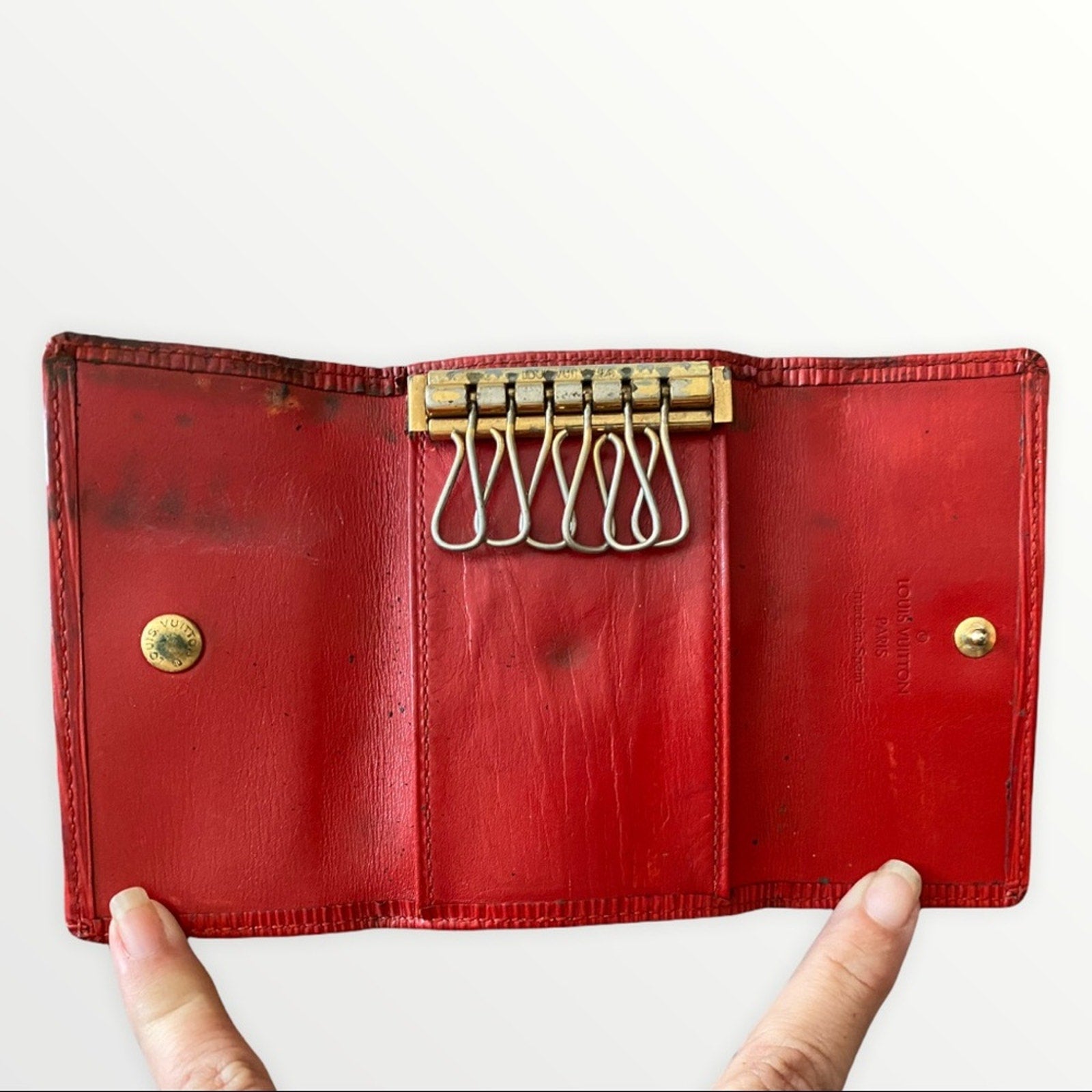 A vintage red leather key ring wallet case held open by two hands against a plain white background. The case features brass hardware, including a key ring mechanism and snap buttons.