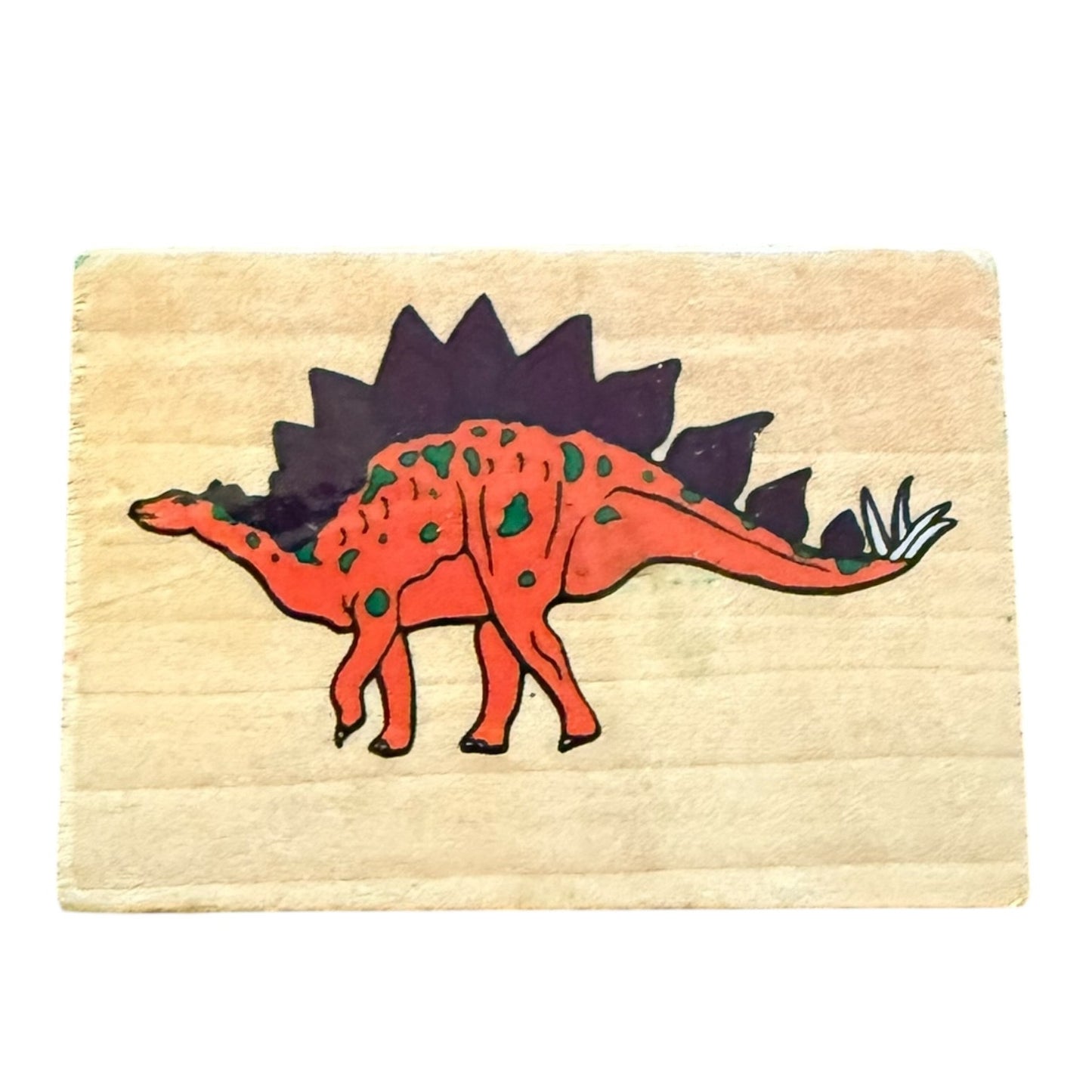 A square wooden stamp featuring a  orange-red Stegosaurus dinosaur with green spots and a dark purple back, on a light wood background.