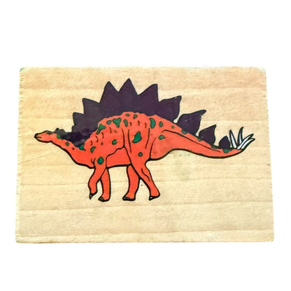 A square wooden stamp featuring a  orange-red Stegosaurus dinosaur with green spots and a dark purple back, on a light wood background.