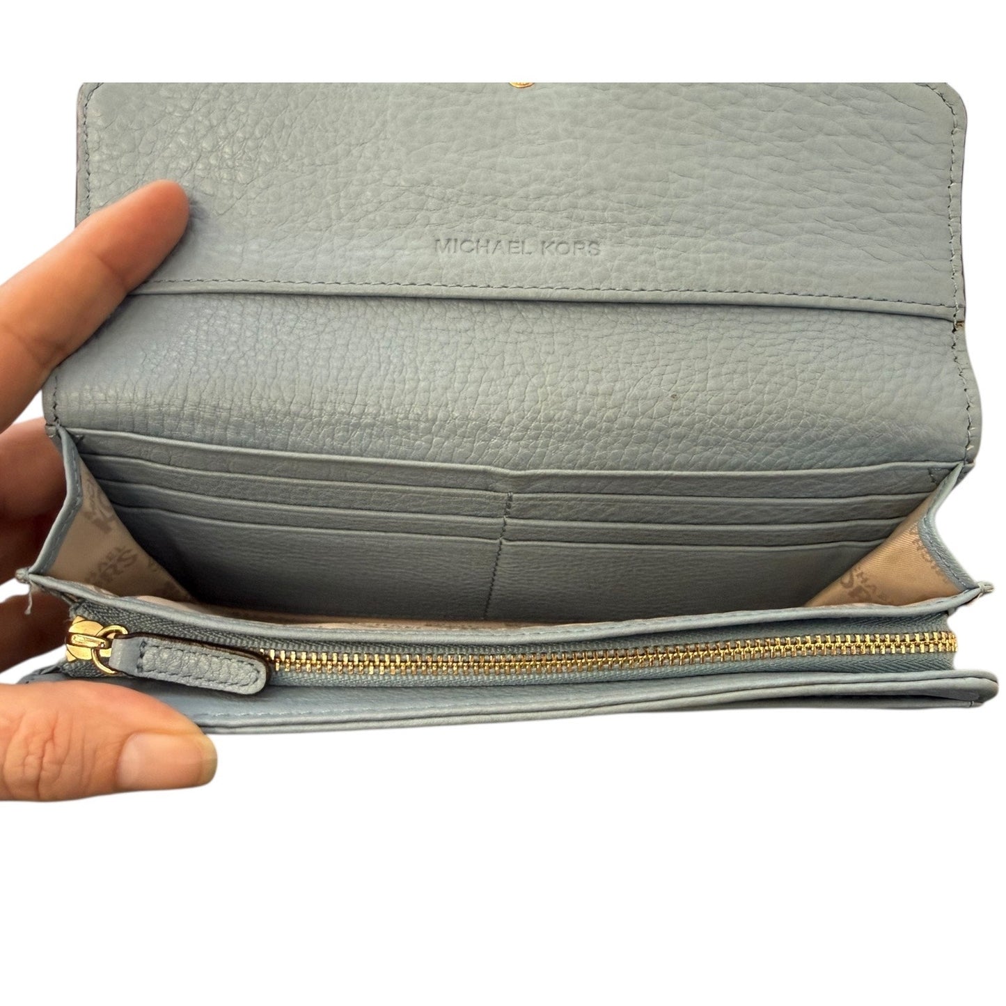 Interior of light blue wallet with gold zipper and pockets held open by a hand, brand 'Michael Kors' visible.