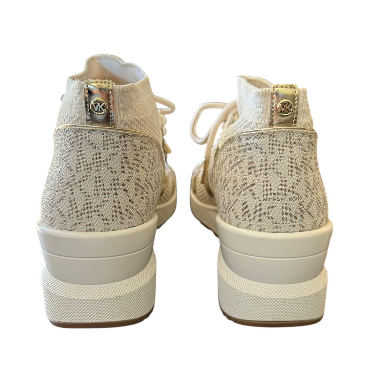 A pair of Michael Kors high-top sneakers shown from the back, featuring a beige and white monogram canvas upper, white laces, and chunky white platform soles with a gold-tone MK logo on the heel tab.