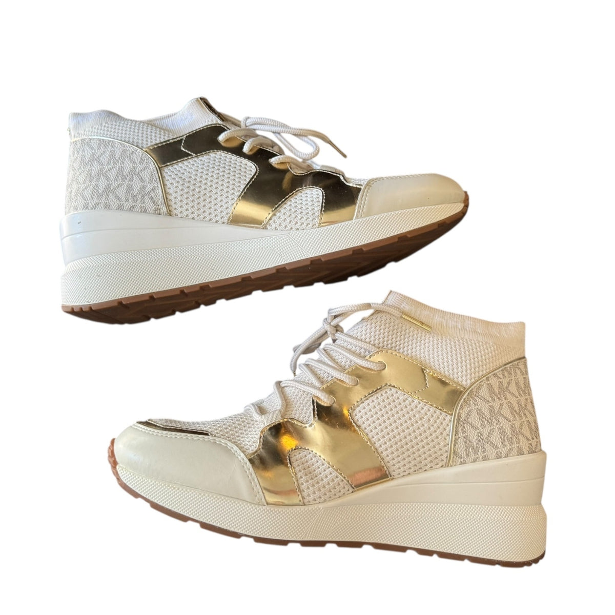A pair of high-top platform sneakers in white and metallic gold, featuring a monogram pattern on the heel, white laces, and a thick white platform sole with a brown outsole.
