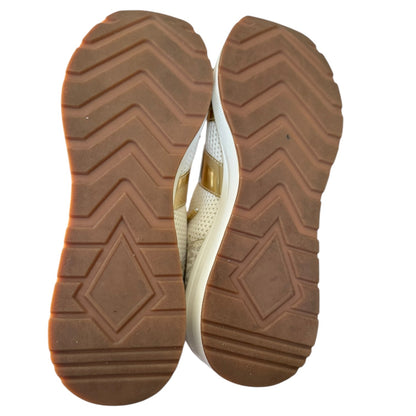 A pair of women's sneakers shown from the sole view, featuring a textured brown chevron-patterned rubber outsole and a white upper with a gold metallic accent.