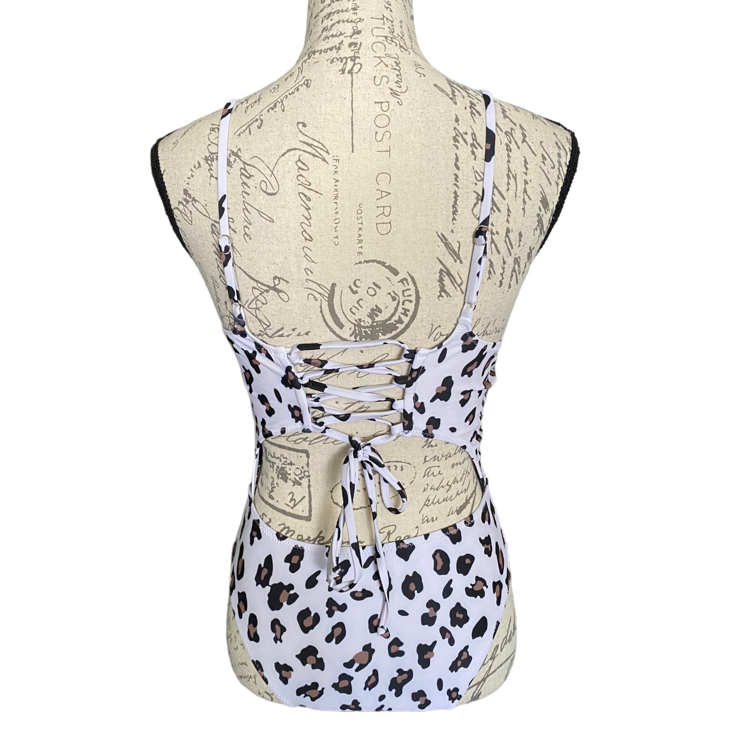A lace-up corset-style back of swimsuit displayed on a fabric dress form mannequin, featuring a white base with black leopard spots and beige vintage postcard print details.