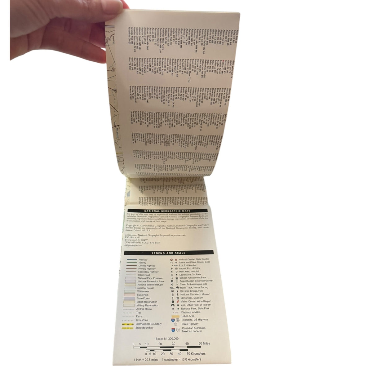 A hand holding open a printed folding map , showing printed text, grid lines, and legend sections on an off-white page against a plain white background.