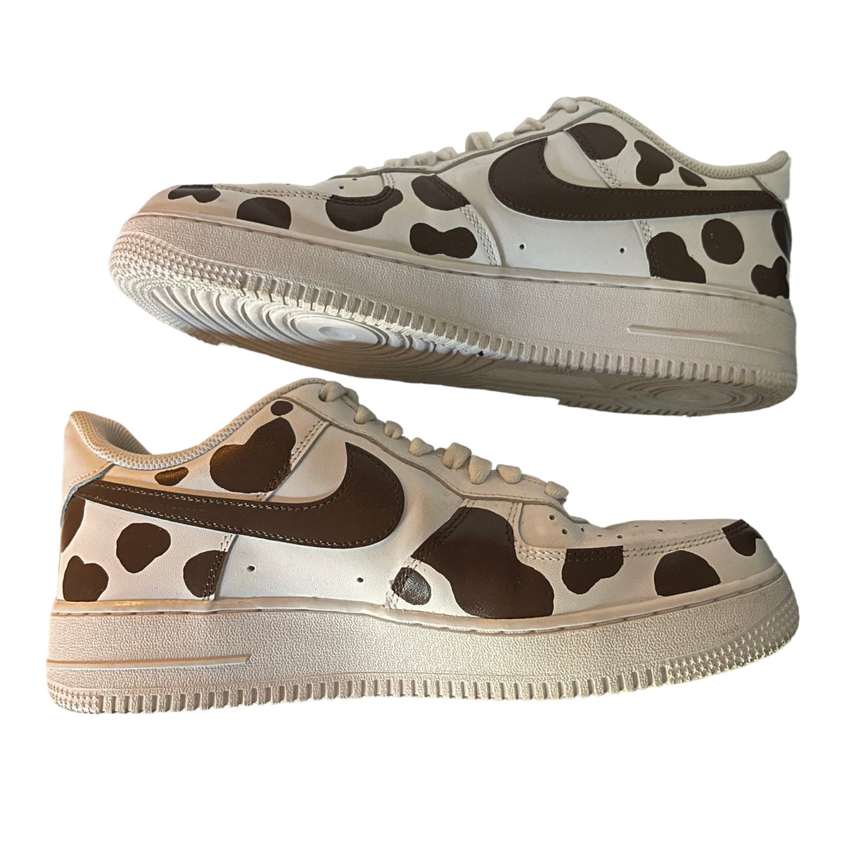 A pair of Nike Air Force 1 Low sneakers featuring a brown cow print pattern on a white base, with dark brown Swooshes.