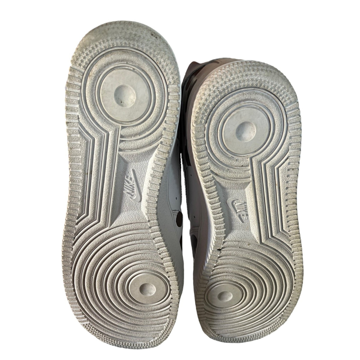 The soles of a pair of Nike sneakers, showing the textured circular tread patterns and embossed Nike logos against a white background.