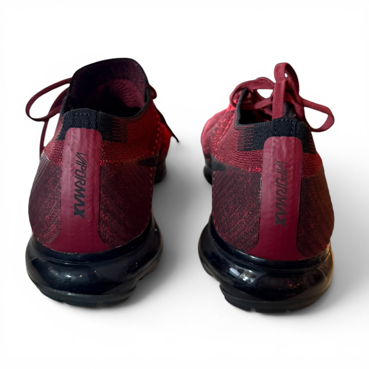 A pair of red and black Nike Air VaporMax running shoes shown from the back, featuring a textured knit upper, burgundy heel tabs with black branding, and black air cushioned soles.