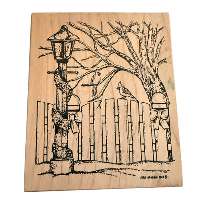 A square wooden block stamp featuring a black illustration of a winter fence scene with a lamppost, bare tree branches, bows, and a small bird, shown on a plain white background.