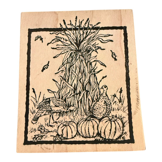 A square wooden stamp block featuring a black ink illustration of autumn harvest elements, including pumpkins, a cornstalk, birds, and leaves, shown on a plain white background.