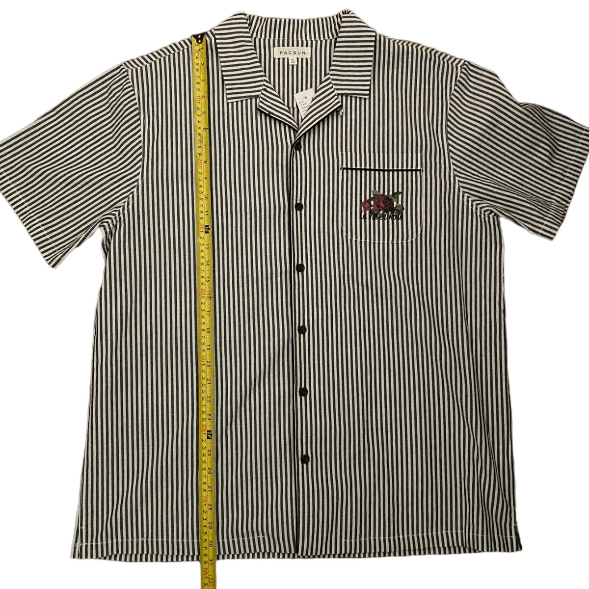 A short-sleeve button-up shirt with vertical black and white stripes, featuring a chest pocket with a small embroidered logo and a yellow measuring tape standing vertically next to it for size reference, set against a plain white background.