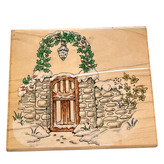 A square wooden board or plaque featuring a painted winter garden scene with a snow-covered stone wall, brown door, green ivy arch, and hanging lantern, photographed on a white background.