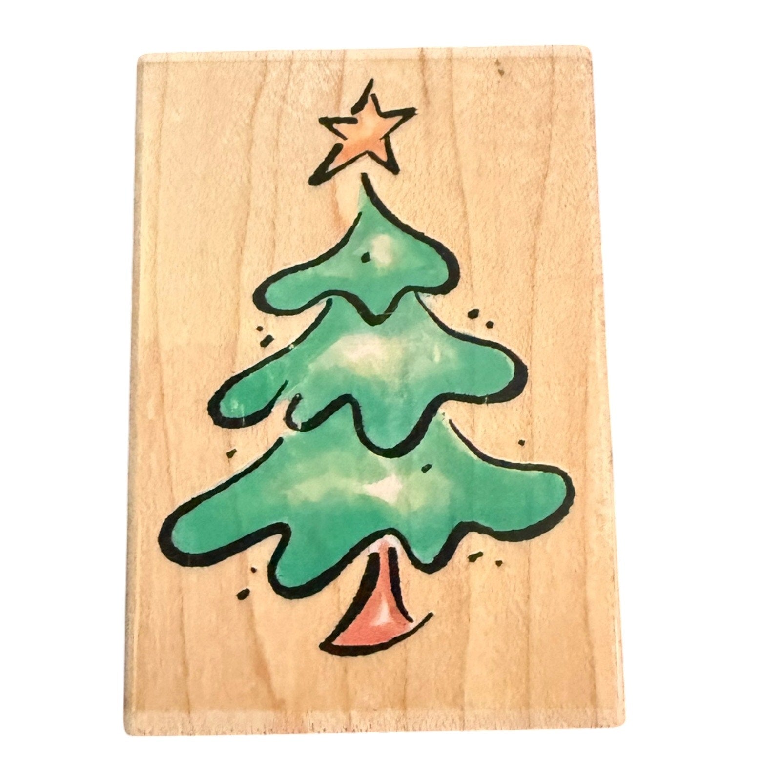 A rectangular wooden block or stamp featuring green Christmas tree with a black outline, a small orange star on top, and an orange bell at the bottom, shown against a plain white background.