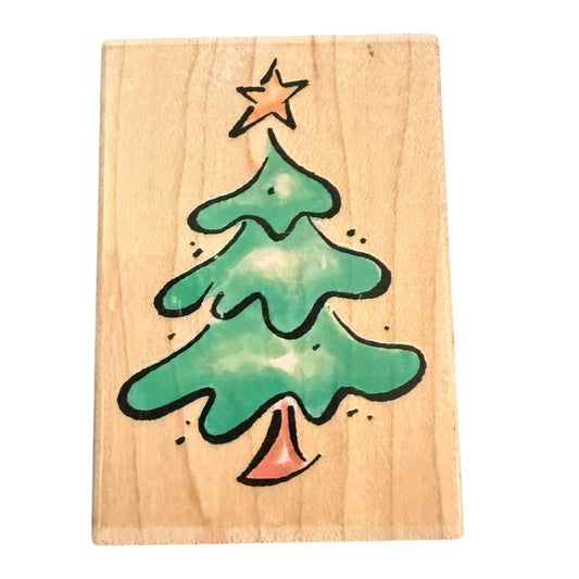 A rectangular wooden block or stamp featuring green Christmas tree with a black outline, a small orange star on top, and an orange bell at the bottom, shown against a plain white background.