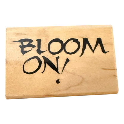 A rectangular wooden stamp with the words 'BLOOM ON!' written in black stylized lettering, shown on a plain white background.