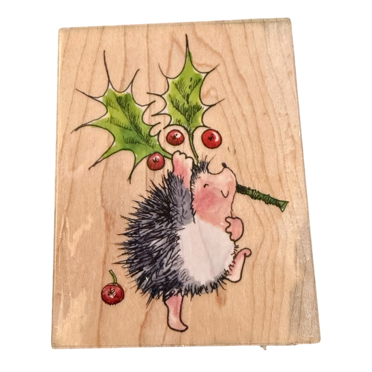 A square wooden block featuring a  illustration of a small hedgehog reaching for green holly leaves with red berries, shown on a plain white background.