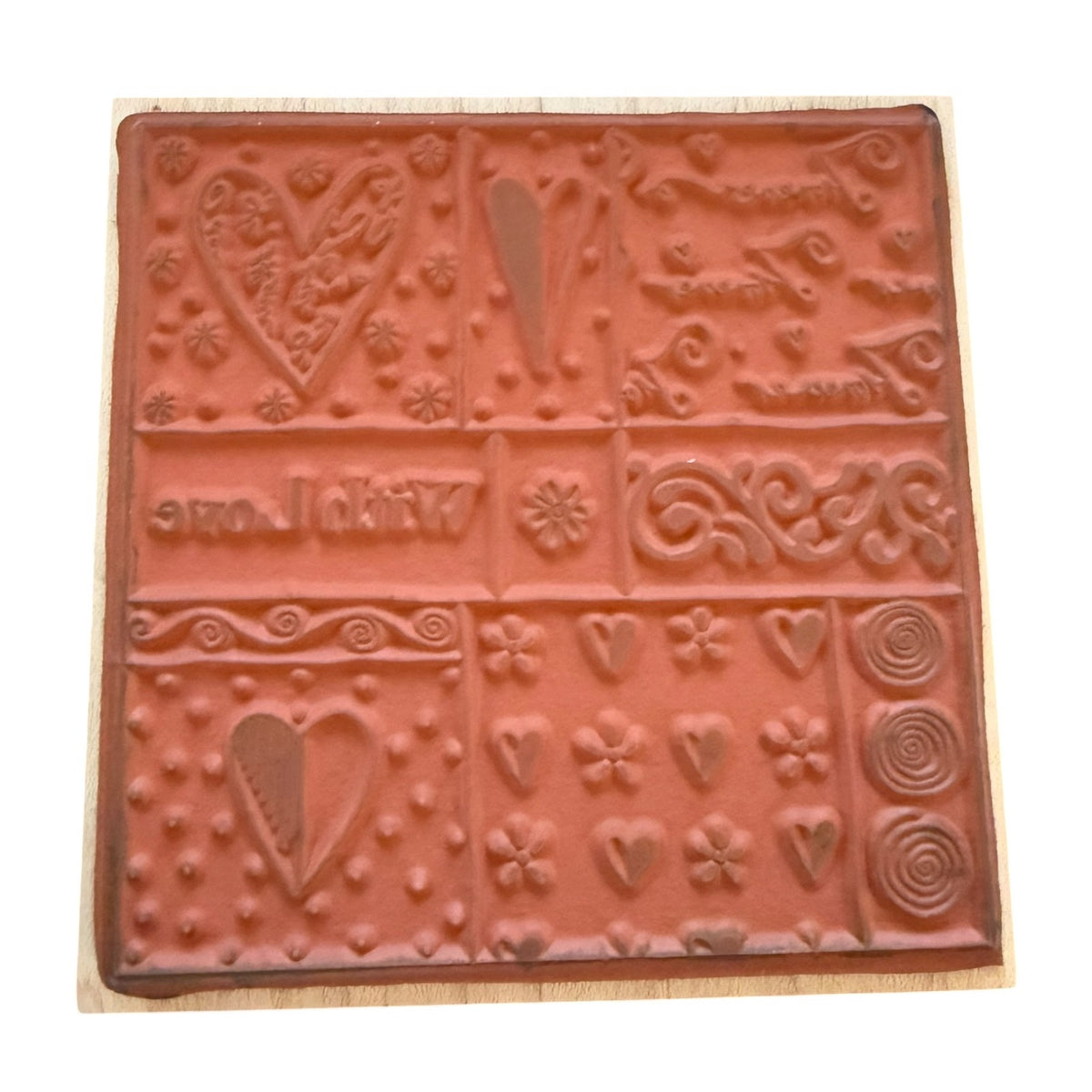 A square terracotta-colored rubber stamp with embossed floral, heart, and scroll designs, mounted on a light-colored wooden block against a white background.