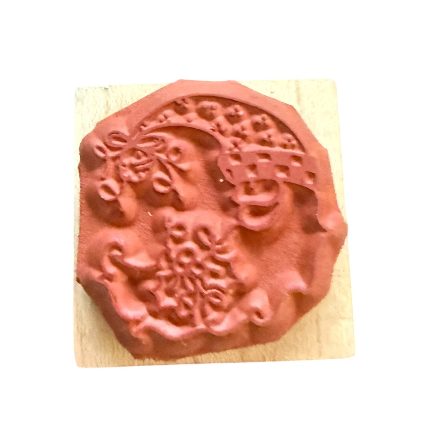 A terracotta-colored rubber stamp with an ornate floral and scroll design, mounted on a light-colored wooden block against a plain white background.