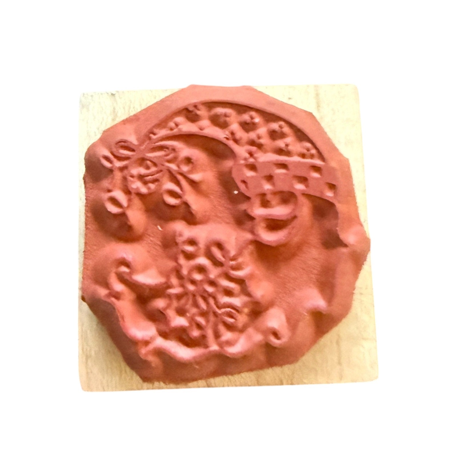 A terracotta-colored rubber stamp with an ornate floral and scroll design, mounted on a light-colored wooden block against a plain white background.