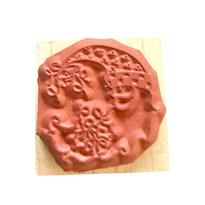 A terracotta-colored rubber stamp with an ornate floral and scroll design, mounted on a light-colored wooden block against a plain white background.