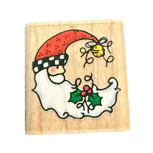 A square wooden stamp block featuring  Christmas design of a white-bearded figure wearing a red hat with a black-and-white check pattern, holding green holly with red berries, and a small yellow bell above.