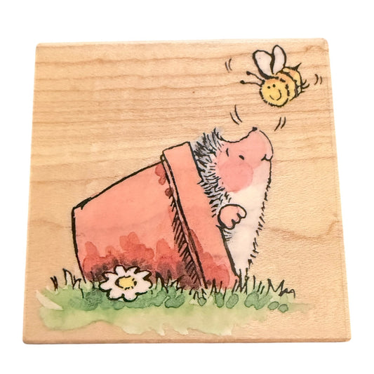 A square wooden block featuring a illustration of a hedgehog peeking out of a tipped-over pink pot, with a yellow bee flying above and a small white flower in green grass below, shown on a plain white background.
