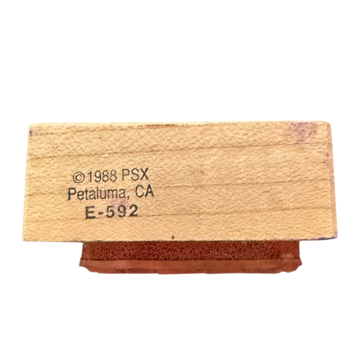 A wooden stamp block with a reddish- rubber stamp head, featuring black printed text that reads '©1988 PSX, Petaluma, CA, E-592' on a plain white background.