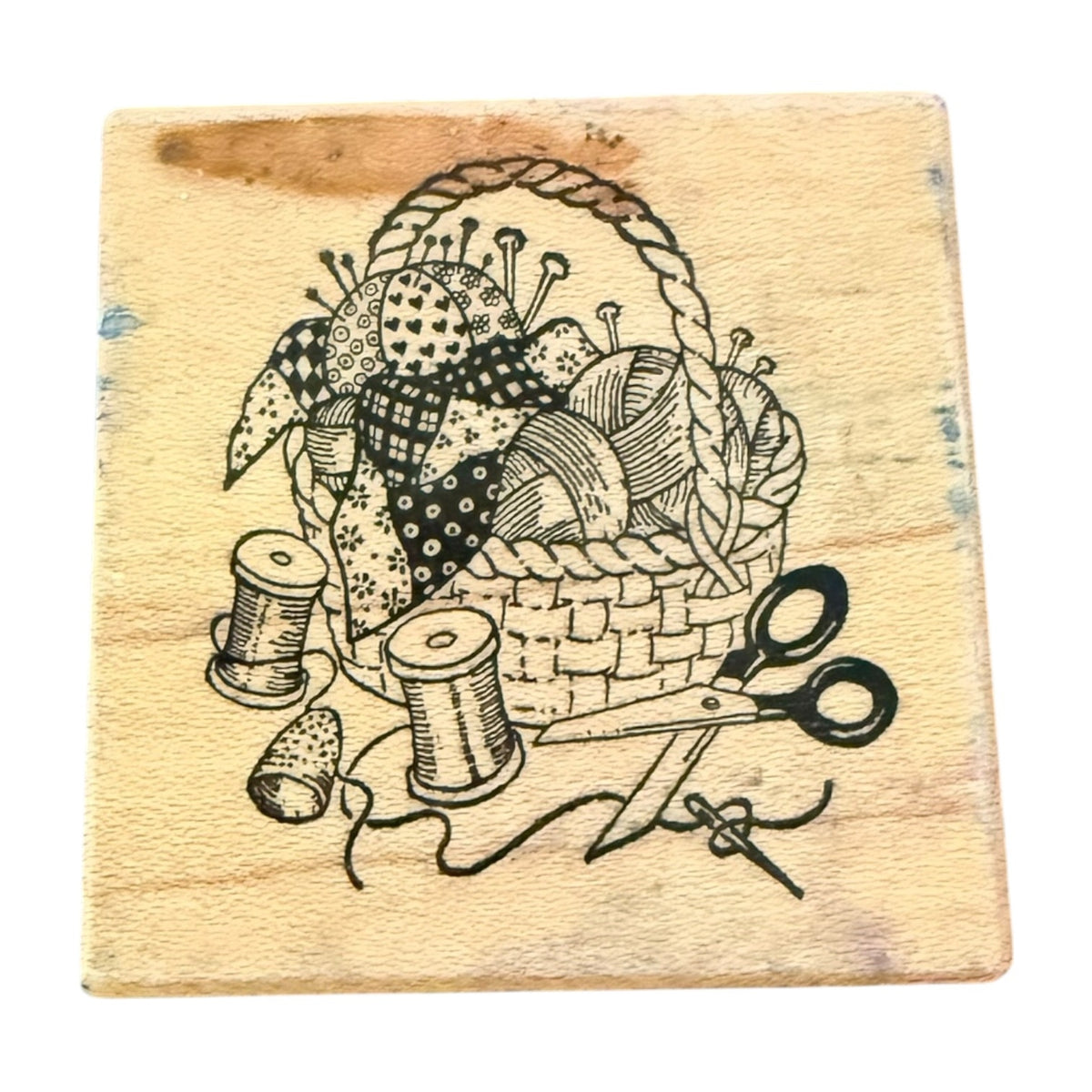 A square wooden block featuring a black printed illustration of a sewing basket with fabric pieces, thread spools, scissors, and a thimble.