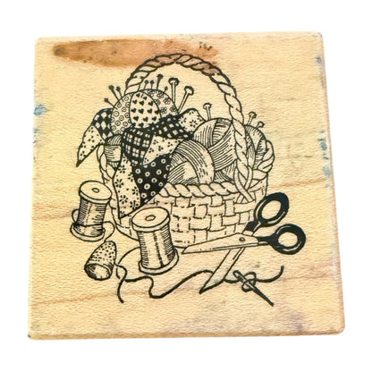 A square wooden block featuring a black printed illustration of a sewing basket with fabric pieces, thread spools, scissors, and a thimble.