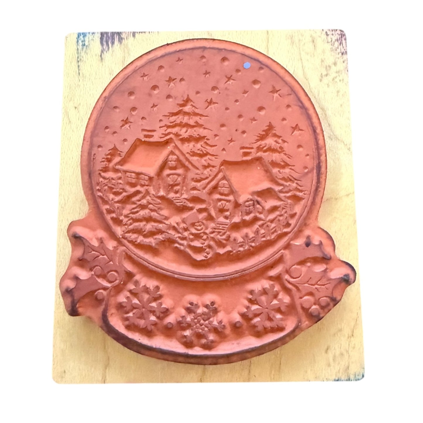 A rubber stamp featuring a winter village scene with houses, trees, and stars in a snow globe, mounted on a light wood block against a white background.