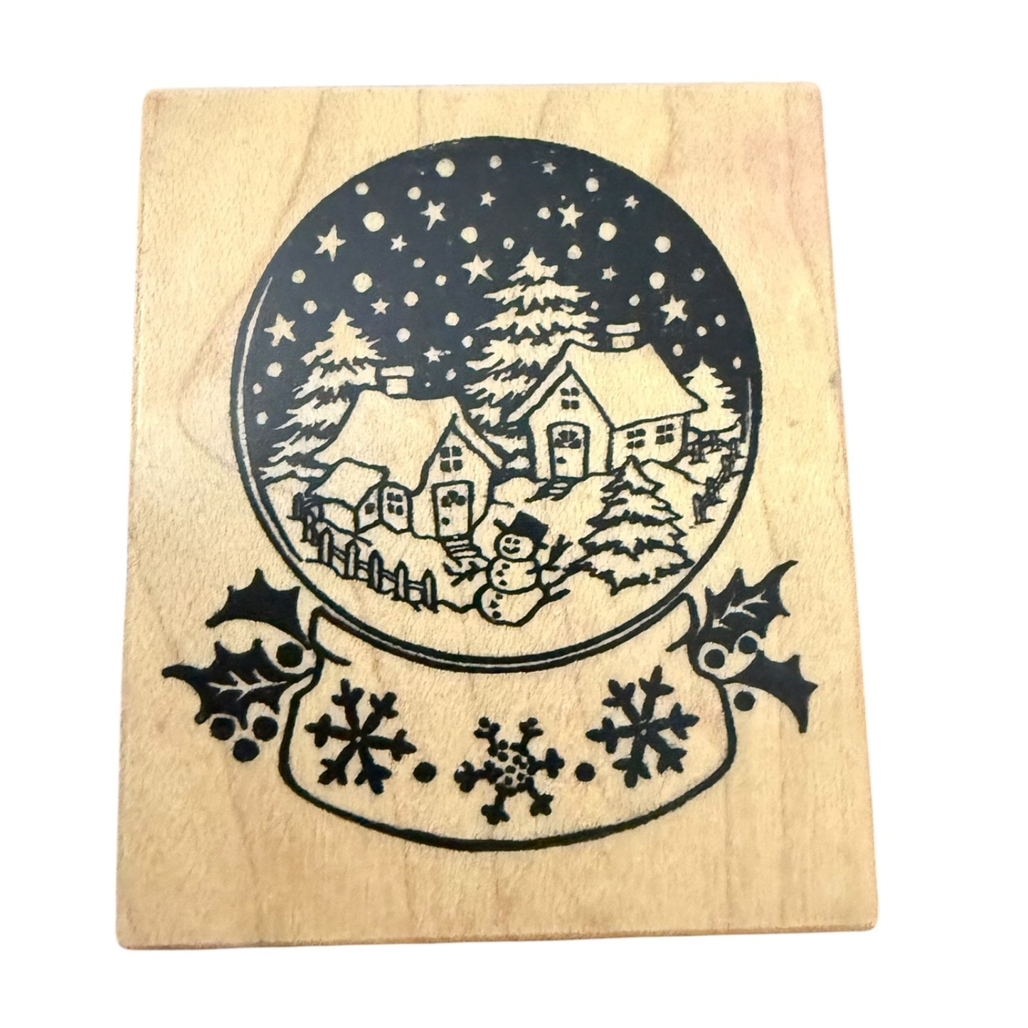 A square wooden stamp block featuring a winter village scene inside a snow globe, with snowflakes and holly on the banner below, shown on a white background.