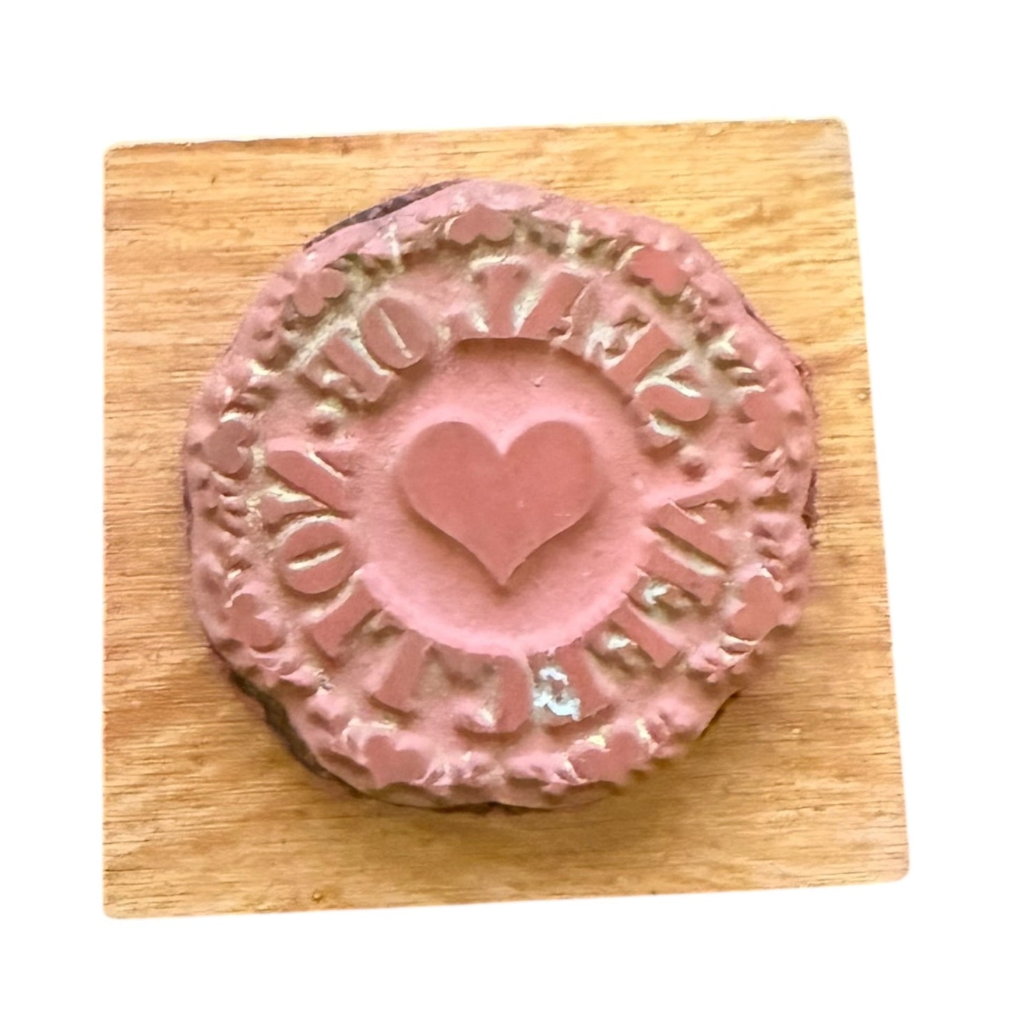 A round pink stamp with a heart in the center and decorative border text, displayed on a square wooden block against a white background.