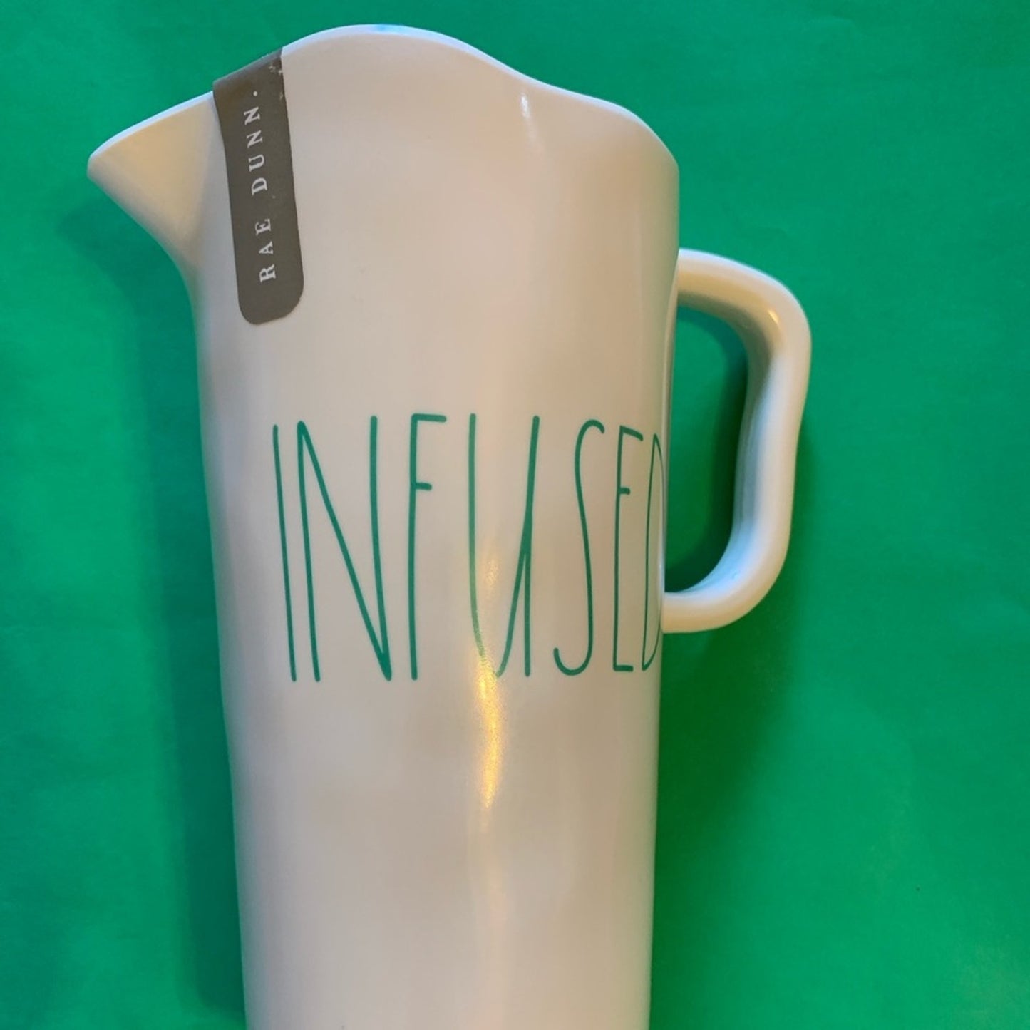 A white pitcher with green text reading 'INFUSED' on the front, a gray tag that says 'RAE DUNN', and a green background.