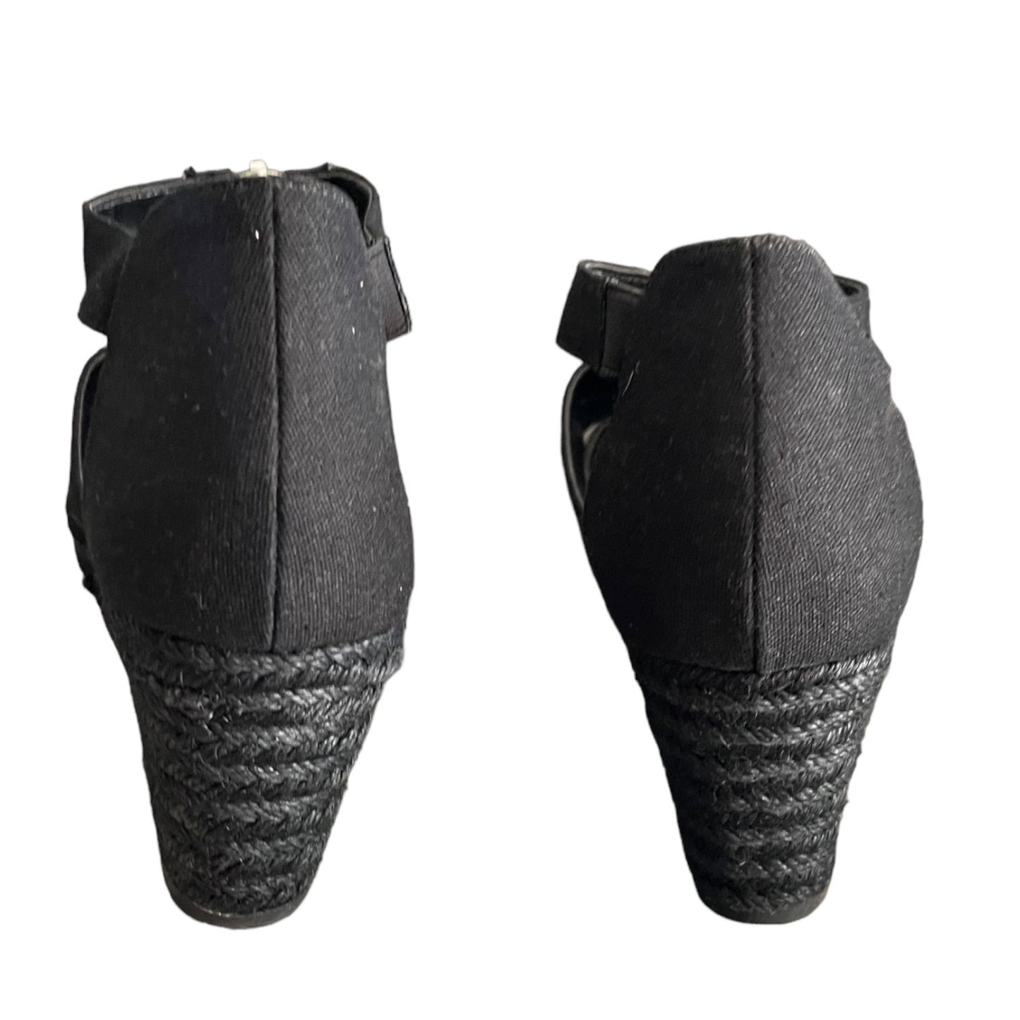 A pair of black wedge sandals shown from the back, featuring a woven jute wedge heel and a black strap with a small metallic buckle.
