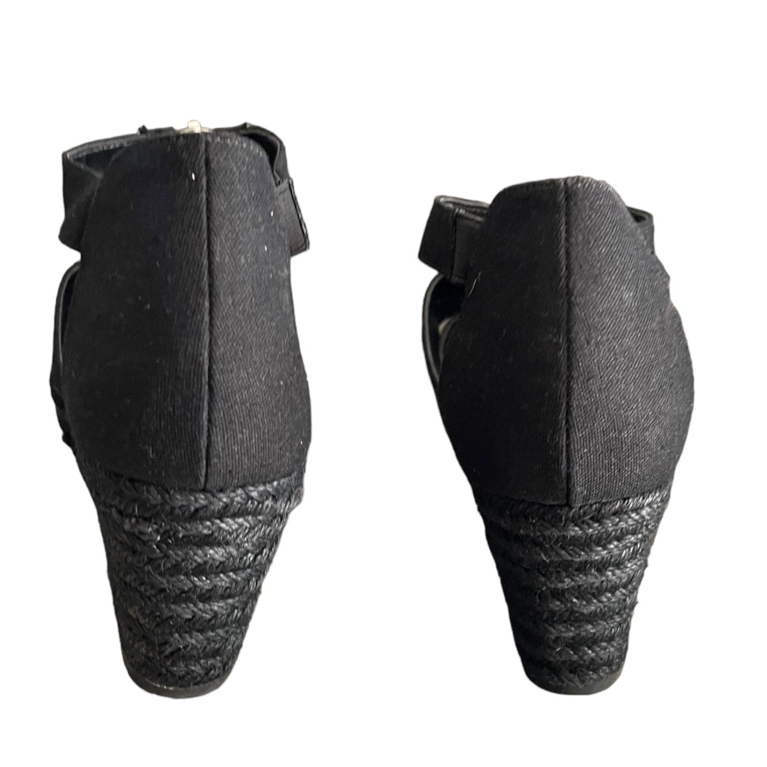 A pair of black wedge sandals shown from the back, featuring a woven jute wedge heel and a black strap with a small metallic buckle.