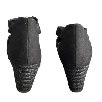 A pair of black wedge sandals shown from the back, featuring a woven jute wedge heel and a black strap with a small metallic buckle.