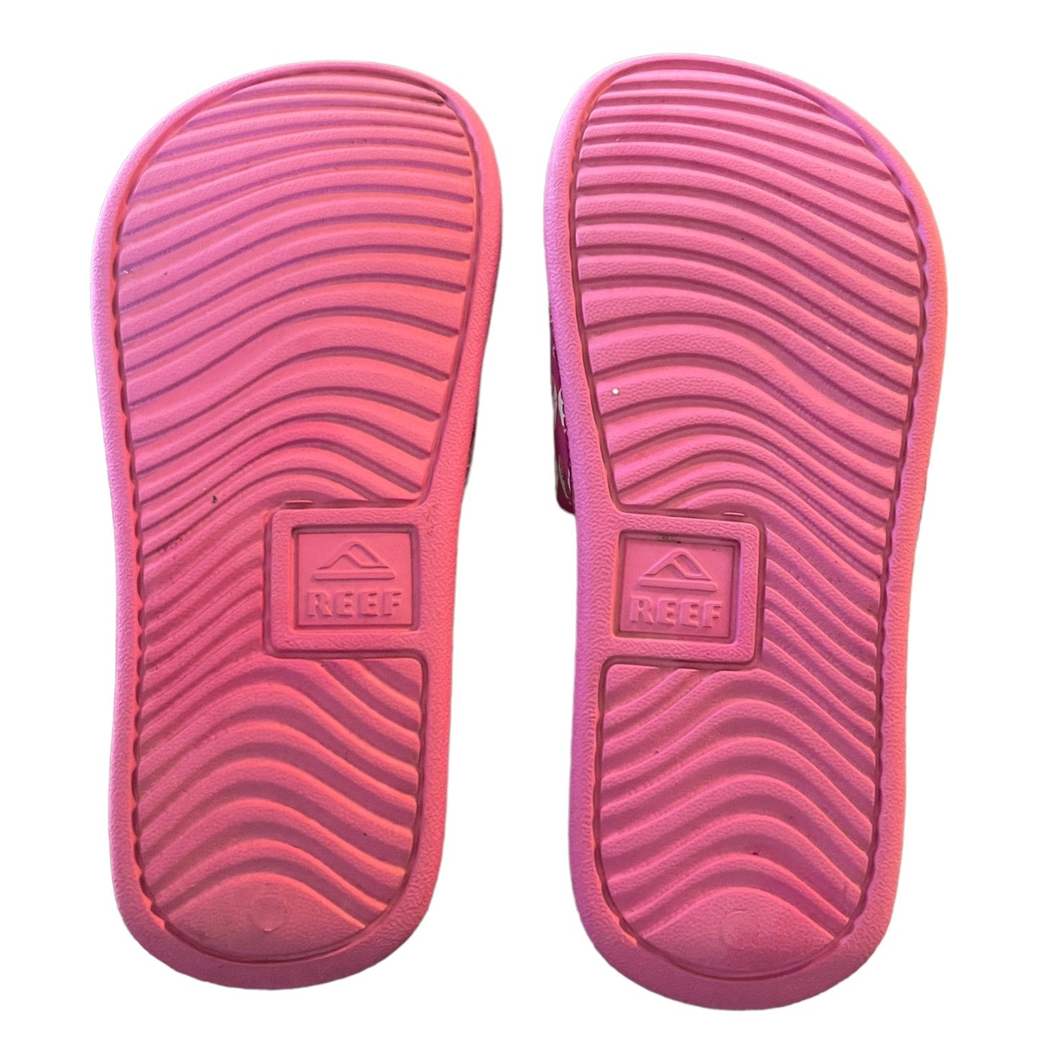 A pair of pink Reef brand slide sandals shown from the sole side, featuring a wavy tread pattern and the embossed Reef logo.