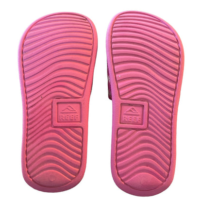 A pair of pink Reef brand slide sandals shown from the sole side, featuring a wavy tread pattern and the embossed Reef logo.