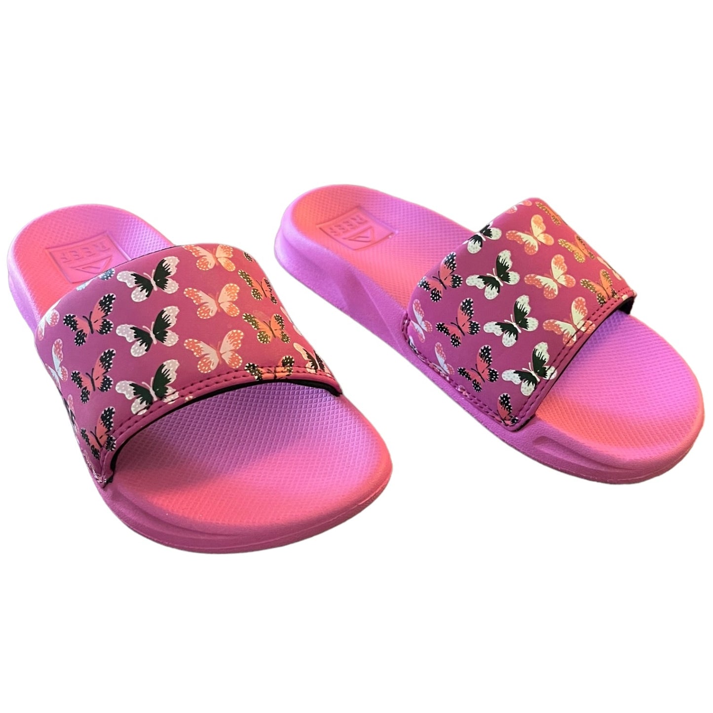 A pair of pink slide sandals with a vibrant pink textured footbed and strap, decorated with a colorful butterfly pattern. The brand logo 'REEF' is visible on the footbed.
