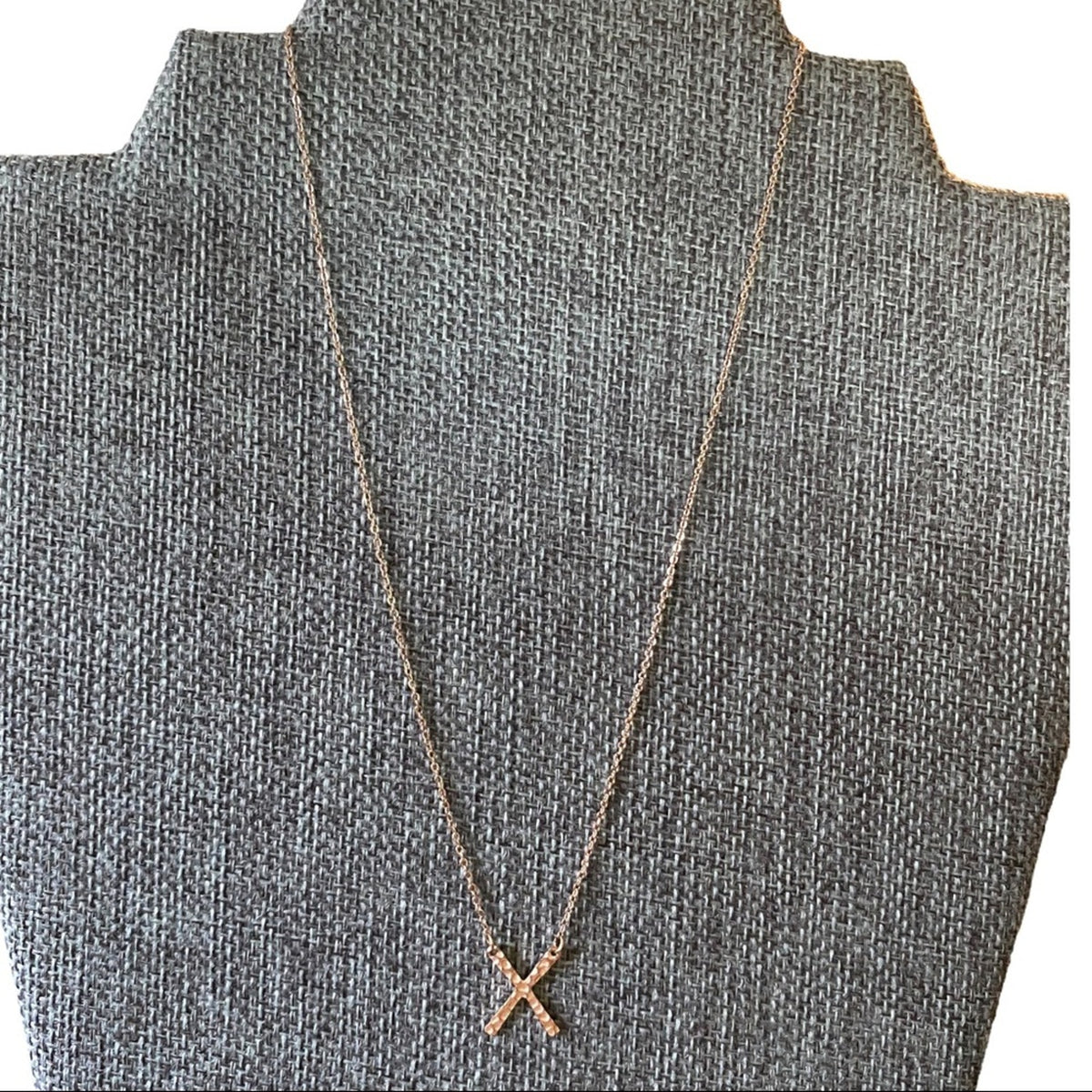A delicate rose gold necklace with an 'X' pendant displayed on a textured gray jewelry bust.